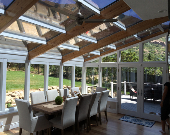 Interior of a straight-eave sunroom with wooden beams and a ceiling fan, white framing, and double doors leading outside, with a long dining table with white high-back chairs around it inside.